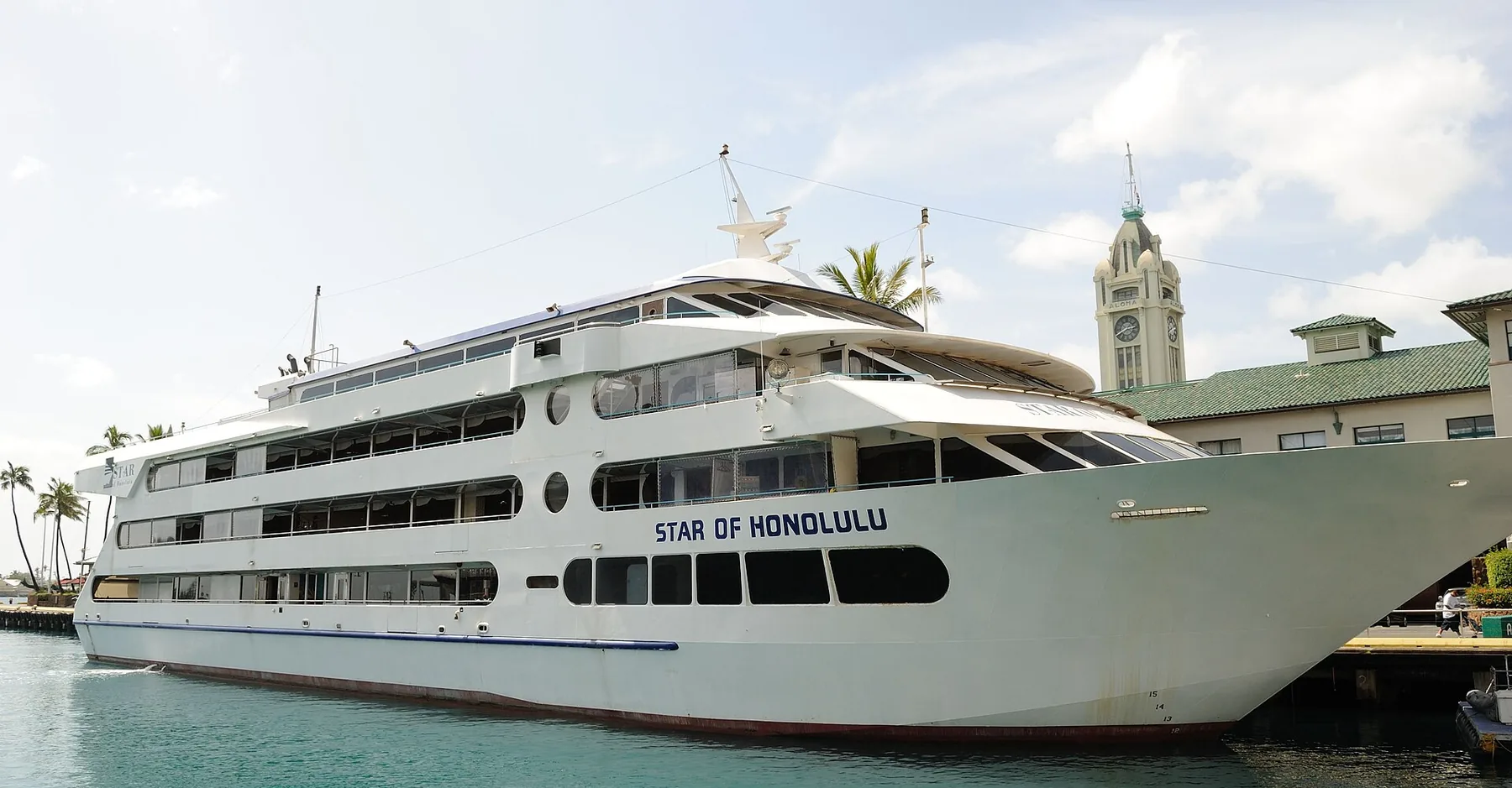The white four-deck Star of Honolulu moored at its Honolulu Harbor pier beside the Aloha Tower clock tower